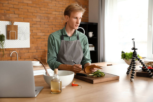 Νεαρός άνδρας κρατώντας σημειώσεις από το μαγείρεμα βίντεο φροντιστήριο στην κουζίνα - Φωτογραφία, εικόνα