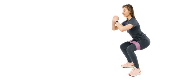 Banner-Foto einer fitten jungen Frau, die mit einem Gummiband Kniebeugen macht. Studioaufnahme vor weißem Hintergrund. - Foto, Bild
