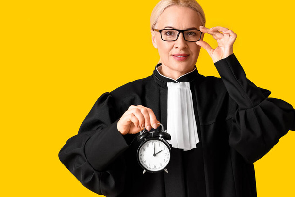 Ώριμη γυναίκα δικαστής με ρόμπα με ξυπνητήρι σε κίτρινο φόντο - Φωτογραφία, εικόνα