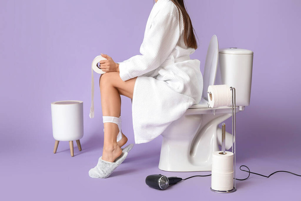 Νεαρή γυναίκα με μπουρνούζι κάθεται σε λεκάνη τουαλέτας με φόντο το χρώμα - Φωτογραφία, εικόνα