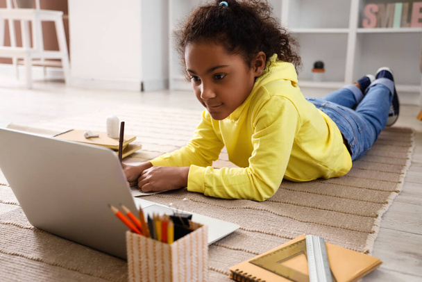 Niña afroamericana escribiendo en copybook mientras estudia con tutor en línea en el piso en casa - Foto, Imagen