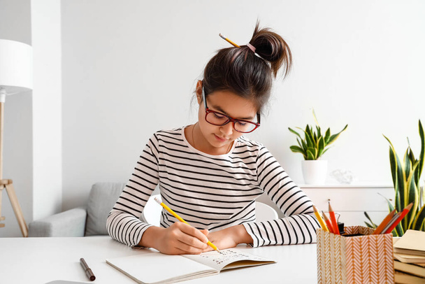 Pequeña chica asiática escribiendo en copybook mientras estudia con tutor en línea en casa - Foto, imagen