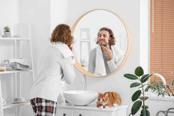 Banyoda dişlerini fırçalayan kedisi olan genç bir adam. - Fotoğraf, Görsel
