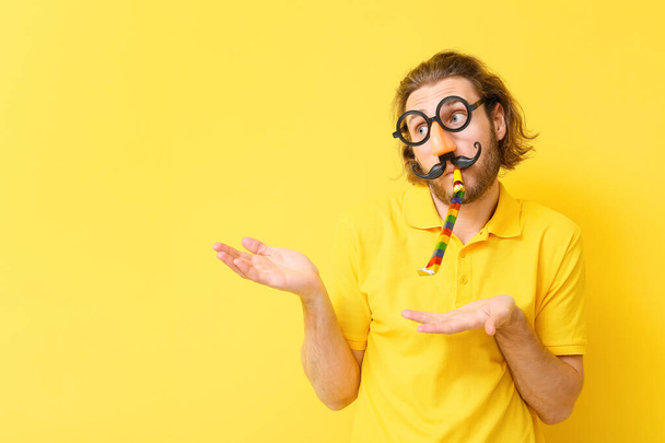 Funny young man in disguise and with party whistle showing something on yellow background. April fools' day celebration - Photo, Image