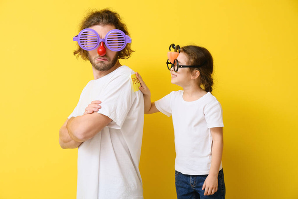 Klein meisje die een grap uithaalt met haar vader tegen een gele achtergrond. April Fools Day viering - Foto, afbeelding