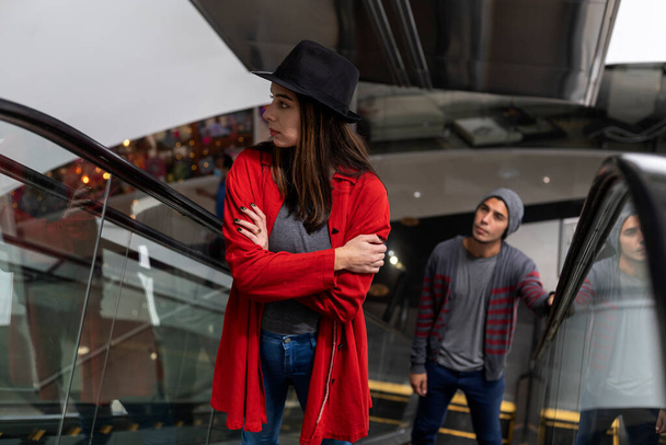 Deux jeunes frères latino-américains montent les escaliers mécaniques dans un centre commercial. Elle porte un chapeau noir et un pull rouge, il porte un chapeau et un pull gris. Concept de marche et de loisirs. - Photo, image