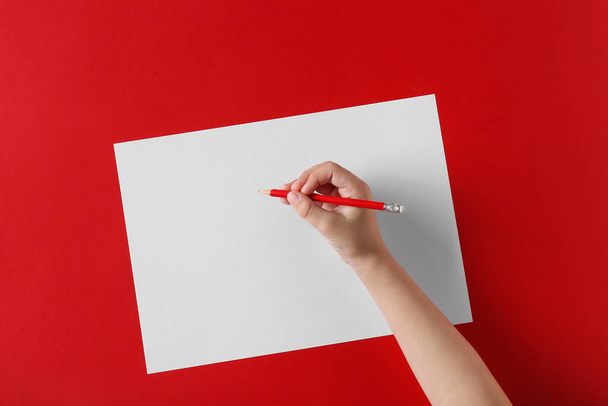 Mano del niño con lápiz y hoja de papel en blanco sobre fondo rojo - Foto, Imagen
