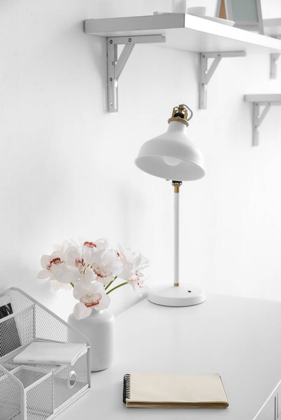 Workplace with modern lamp, notebook and vase with flowers near white wall - Photo, Image