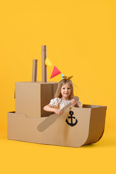 Cute little girl playing with cardboard ship on yellow background - Foto, afbeelding