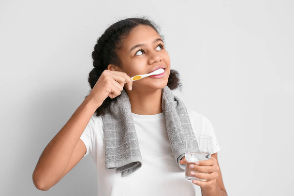 Afro-Amerikaanse tiener meisje met glas water poetsen tanden op lichte achtergrond - Foto, afbeelding