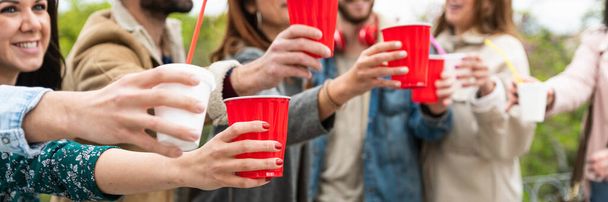 νέοι άνθρωποι που διασκεδάζουν παράδοση πίνοντας καφέ μαζί χάρτινο φλιτζάνι παρέα ποτό σε πλαστικό κύπελλο στο χέρι closeup χέρια των φίλων ψήσιμο με κόκκινο πλαστικό κύπελλο - οριζόντια banner ή κεφαλίδα - Φωτογραφία, εικόνα