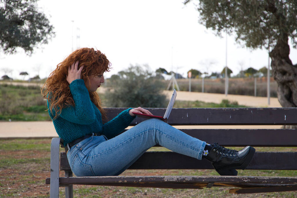 Genç ve güzel kızıl saçlı kadın birçok zeytin ağacının olduğu bir parkta bir bankta oturuyor. Dizüstü bilgisayarında sosyal ağları kontrol ediyor ve etkili olarak çalışıyor. Öğrenci kavramı. - Fotoğraf, Görsel