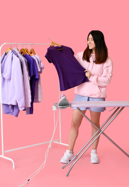 Mujer asiática joven con camiseta, tabla de planchar y estante sobre fondo rosa - Foto, Imagen