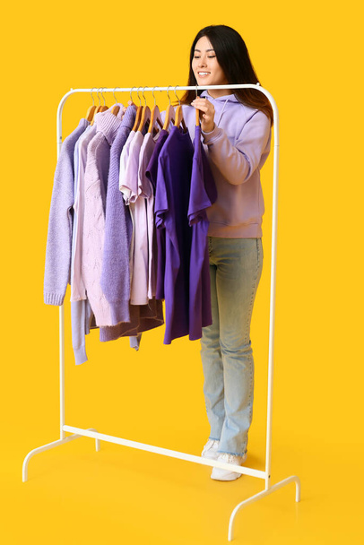 Jeune femme asiatique et rack avec des vêtements dans les tons violets sur fond jaune - Photo, image