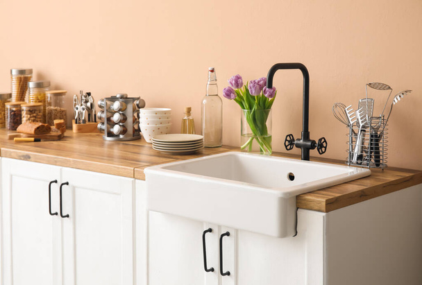 Bílé pulty s umyvadlem, tulipány ve váze a kuchyňské nádobí u béžové stěny - Fotografie, Obrázek