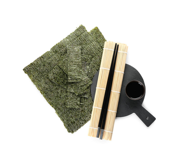 Доска с палочками для еды, бамбуковым ковриком, соевым соусом и вкусными простынями из водорослей на белом фоне - Фото, изображение