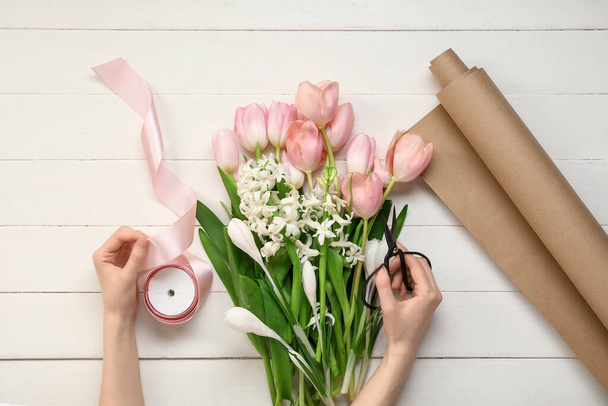 Female hands with beautiful spring flowers, scissors and ribbon on light wooden background - Fotografie, Obrázek