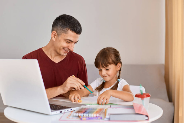 Indoor shot of smiling loving young father sitting with schoolgirl daughter at table and helping doing homework, tutor coming to teach a cute schoolgirl. - Fotoğraf, Görsel