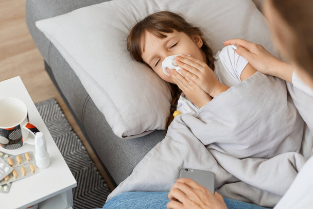 Horizontal shot of ill sick little girl lying on sofa under blanket and sneezing, faceless mother sitting near unhealthy daughter, virus, influenza symptoms. - Фото, изображение