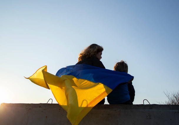 niño y una mujer se sientan de espaldas al atardecer contra el cielo con una bandera azul y amarilla ucraniana. Familia, refugiados, unidad, apoyo. Los ucranianos están en contra de la guerra. de pie con Ucrania - Foto, imagen