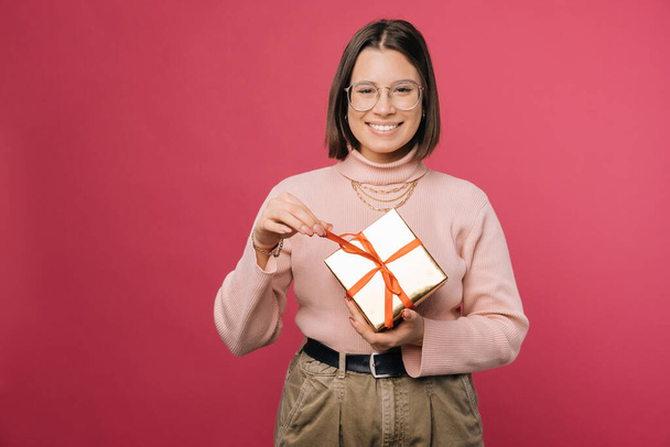 Joyful woman is unwrapping a gift box while smiling at the camera. Studio shot over pink background. - Foto, imagen