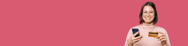 Фотография Панорамы с копией молодой женщины с помощью телефона и кредитной карты. Студия на розовом фоне. - Фото, изображение