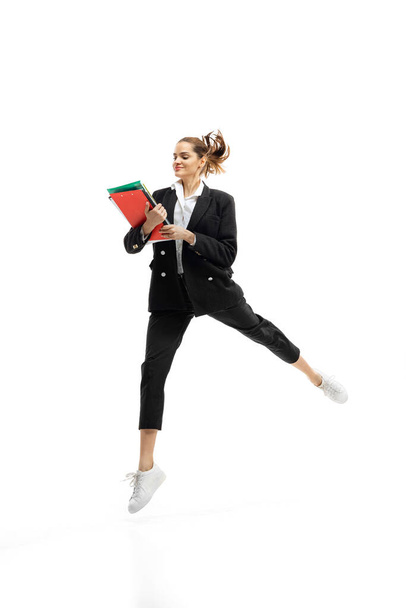 Dynamic portrait of young businesswoman in black suit jumping with folders isolated over white studio background - Foto, imagen