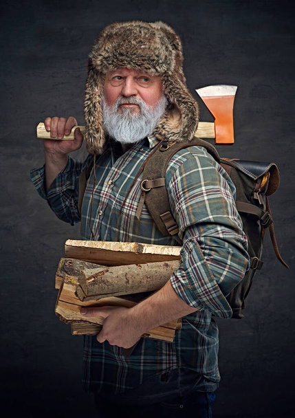 Anciano leñador con sombrero ruso sosteniendo hacha y pila de madera - Foto, Imagen