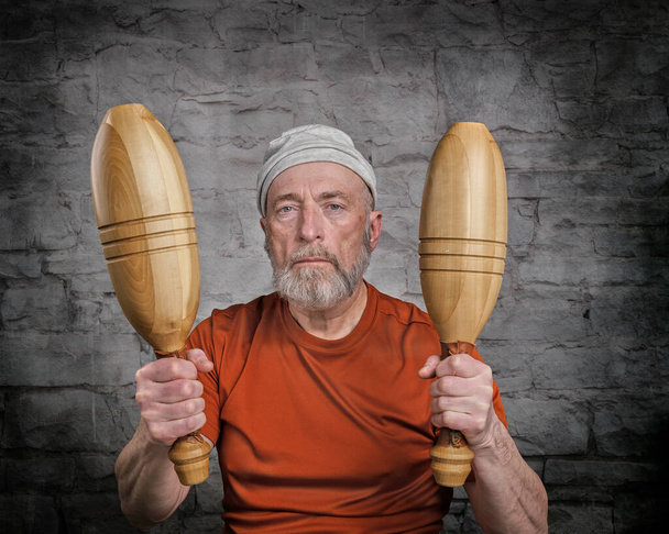 senior man (in late 60s) is exercising with wooden Indian clubs, portrait against dark grunge background, fitness over 60 concept - 写真・画像