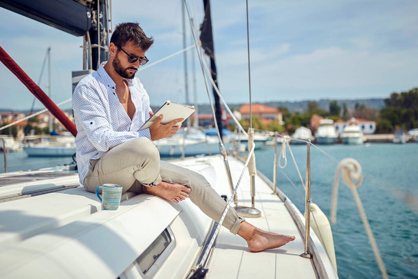 Młody człowiek oglądający zawartość tabletu siedząc na jachcie i jadąc przez dok w piękny dzień nad morzem. Lato, morze, wakacje - Zdjęcie, obraz