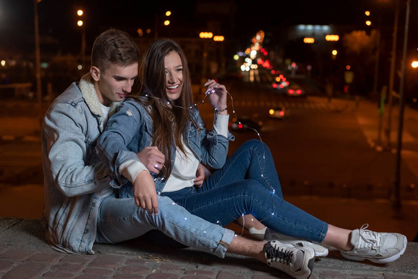 Jeune couple caucasien est assis sur le trottoir embrassant dans le contexte des lumières de la ville de la nuit. - Photo, image