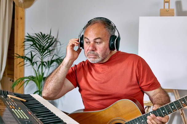 Mature man wearing headphones creating new song while playing acoustic guitar, musical keyboard or synthesizer at home. Music hobby. Creative leisure. Self expression concept. Home music studio. - Fotó, kép