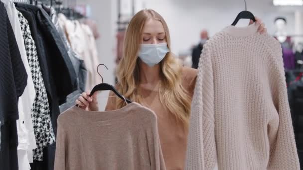 Молодая девушка в медицинской маске выбрать одежду в магазине женщина потребитель сделать выбор между двумя свитерами собирает новый модный образ выбирая прыгунов думая сомнения леди покупки во время пандемии covid19 - Кадры, видео