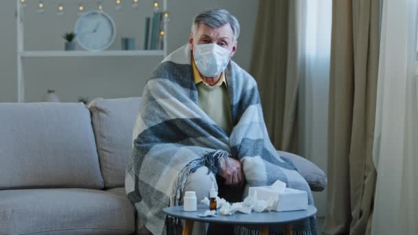 Aufgebrachter kranker Opa in medizinischer Maske sitzt auf Couch kaukasischer älterer Mann in Decke gehüllt hat Schüttelfrost kranker reifer Senior blickt auf Kamera Symptome covid 19 epidemischen Coronavirus Ausbruch Grippe-Konzept - Filmmaterial, Video
