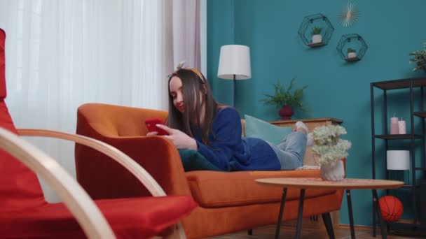 Relaxált mosolygós fehér fiatal nő feküdt otthon kanapén okostelefon görgetés, böngészés online - Felvétel, videó
