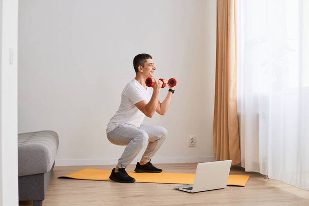 Крытый снимок спортивного спортсмена в спортивной одежде, занимающегося спортивными упражнениями дома, сидящего на корточках, сидящего дома, сосредоточенного взгляда, держащего гантели в руках, - Фото, изображение