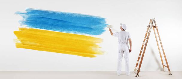 Ukrayna bayrağının sarı ve mavi renklerini boyayan ressam Ukrayna 'da barış ve savaşı durdurma kavramı. Beyaz duvarda izole edilmiş - Fotoğraf, Görsel