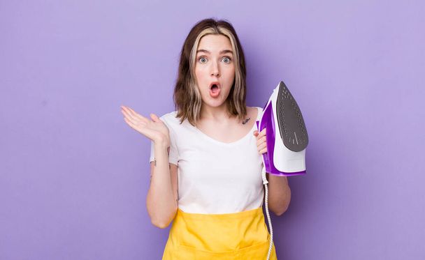 pretty caucasian woman looking surprised and shocked, with jaw dropped holding an object. hand iron concept - Photo, Image