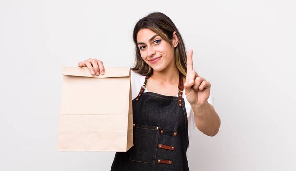 pretty hispanic woman smiling and looking friendly, showing number one. delivery and take away concept - Foto, afbeelding