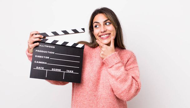 pretty hispanic woman smiling with a happy, confident expression with hand on chin. cinema clapper concept - Foto, imagen