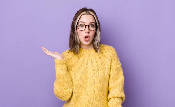pretty caucasian woman looking surprised and shocked, with jaw dropped holding an object with an open hand on the side - Photo, Image