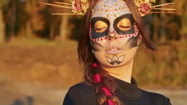 Молодая женщина с макияжем Санта-Муэрте, одетая в черное платье смерти, идет на фоне осенних листьев в лесу на закате. День мертвых, или Хэллоуин. - Кадры, видео