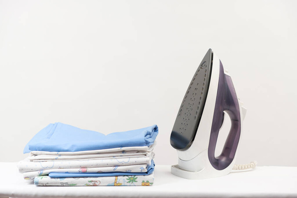 Σιδερένιο και μια στοίβα σιδερωμένα ρούχα και πάνες για μωρά σε λευκό φόντο με χώρο για κείμενο - Φωτογραφία, εικόνα