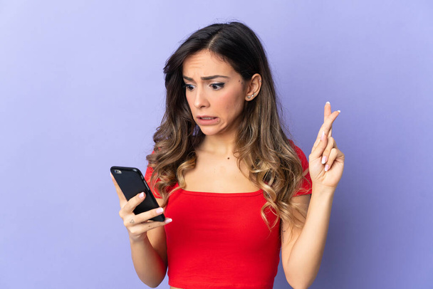 Mladá běloška žena izolované na fialovém pozadí pomocí mobilního telefonu s prsty kříž - Fotografie, Obrázek