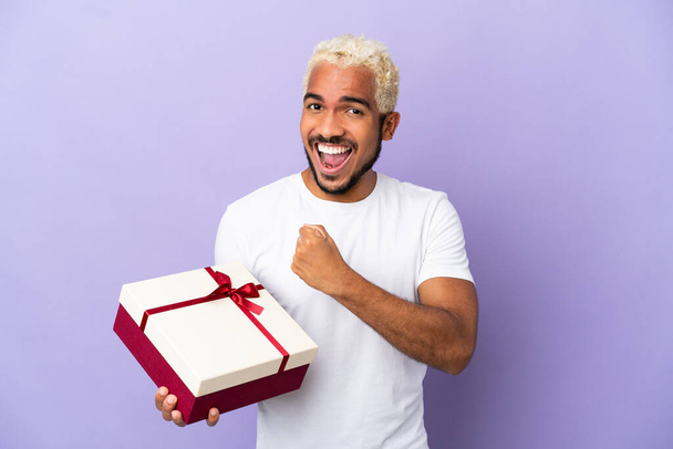 Young Colombian man holding a gift isolated on purple background celebrating a victory - Valokuva, kuva
