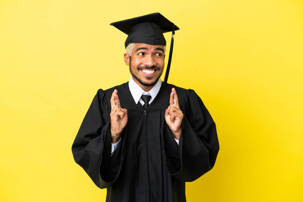 Νεαρός απόφοιτος πανεπιστημίου Κολομβιανός άνδρας απομονωμένος σε κίτρινο φόντο με σταυρωμένα δάχτυλα - Φωτογραφία, εικόνα