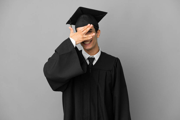Mladý argentinský univerzitní absolvent izolovaný na šedém pozadí zakrývající oči rukama a s úsměvem - Fotografie, Obrázek