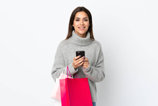 Young caucasian woman isolated on white background holding shopping bags and writing a message with her cell phone to a friend - Zdjęcie, obraz