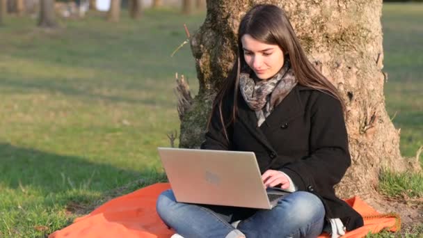 Kaunis nuori brunette nainen työskentelee kannettavan istuu jalka puun puistossa. Hän katselee joskus ympärilleen ja hymyilee kirjoittaessaan näppäimistölle. Etätyökäsite. Kopioi tila. - Materiaali, video