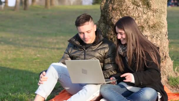 Krásný pár v neformálním oblečení se nadchne při používání notebooku venku. Sedíte na úpatí stromu a vychutnáváte si přírodu za slunečného jarního dne. Koncepce vzdálené týmové práce a pozitivity. - Záběry, video